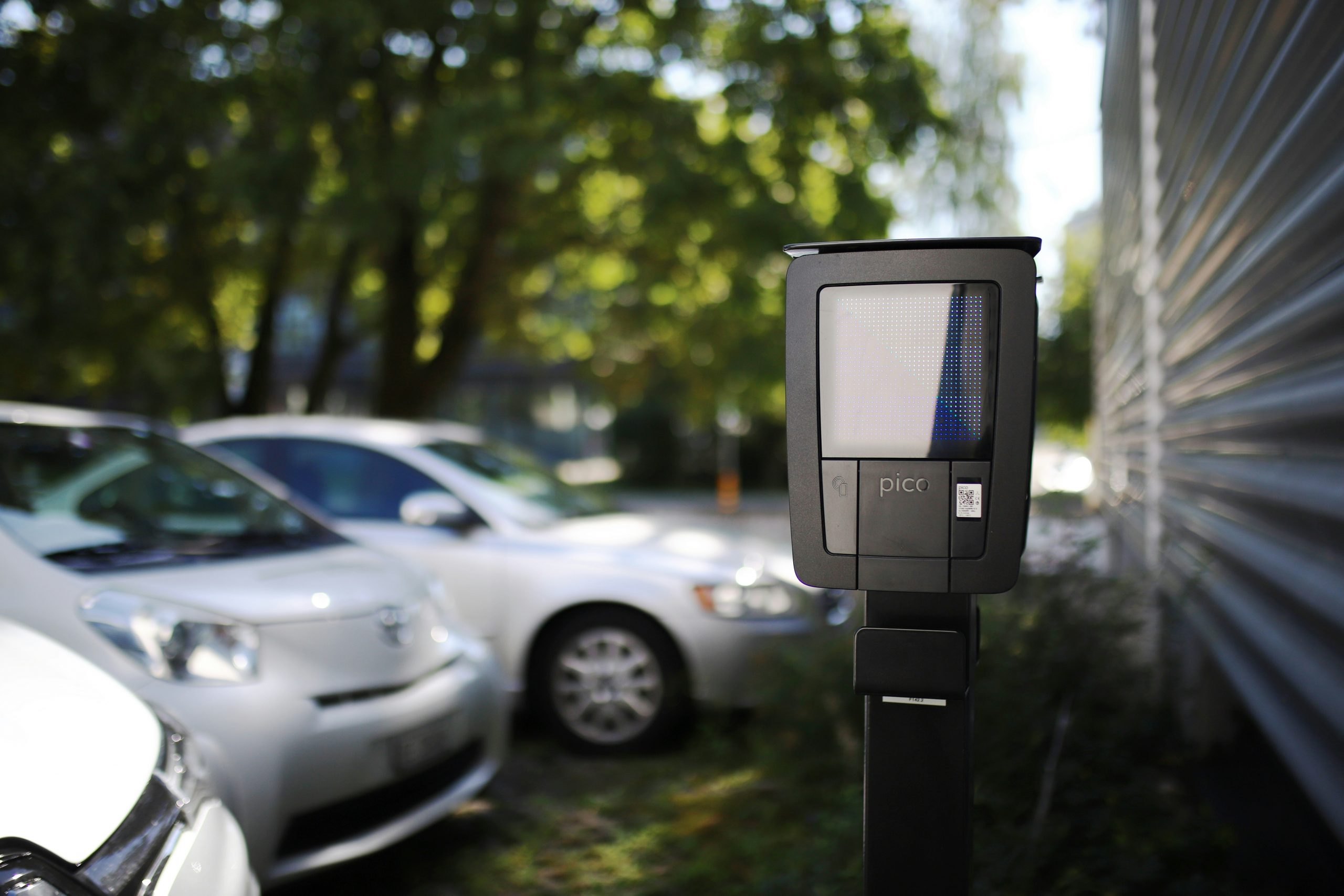 Nahaufnahme einer smarten Ladestation für Elektroautos auf einem Parkplatz mit mehreren geparkten Fahrzeugen im Hintergrund.